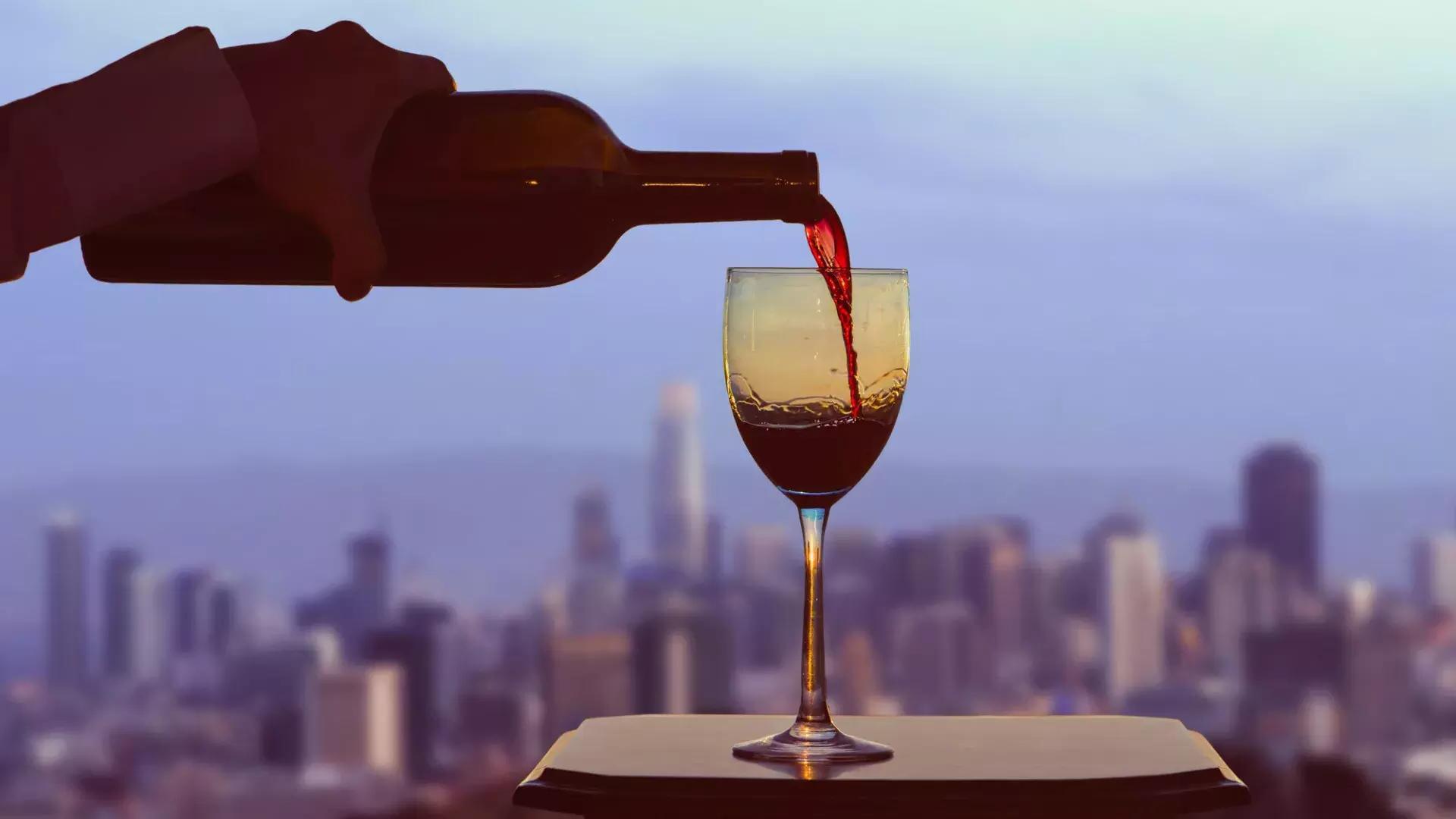 Uma taça de vinho tinto sendo servida, com o horizonte de São Francisco visível pela janela.