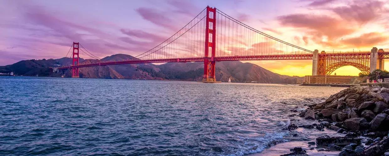 的 金门大桥 at sunset with a multicolored sky and the San Francisco Bay 在 foreground.